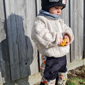 Op de afbeelding: Een kind draagt een zwarte gebreide muts met een wit logo met de tekst "Eisbär" en een rode "S" aan de zijkant. Het kind draagt een crèmekleurige trui en een zwarte corduroy broek met een kleurrijke print van bouwvoertuigen op de benen. Het kind houdt een geel speelgoed vast.