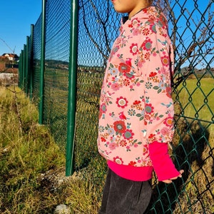 May include: A young girl wearing a pink floral print hoodie with a bright pink zipper and cuffs, black jeans, and white sneakers. She is wearing pink sunglasses and has her hair pulled back with a blue scrunchie.