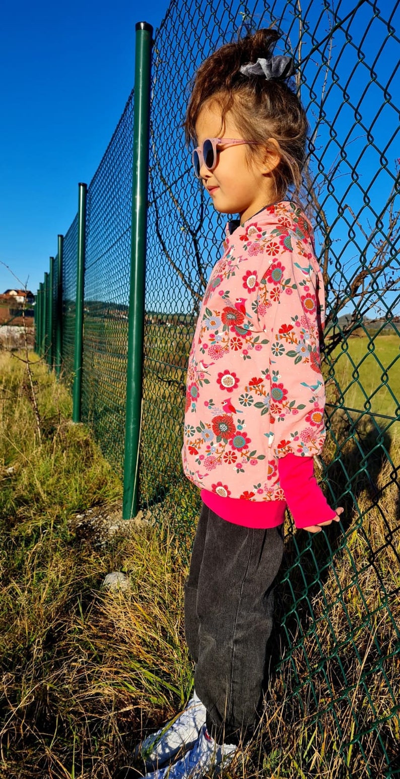 May include: A young girl wearing a pink floral print hoodie with a bright pink zipper and cuffs, black jeans, and white sneakers. She is wearing pink sunglasses and has her hair pulled back with a blue scrunchie.