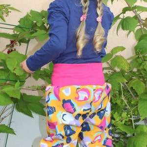 May include: A young girl wearing a blue long-sleeved shirt with a pink waistband and colorful patterned pants. She has her blonde hair in two braids with pink hair clips.