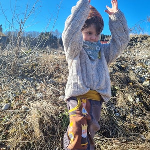 May include: A child wearing a light brown sweater with a white knit pattern, a brown and orange knit hat, and a green bandana with a white floral pattern. The child is wearing brown corduroy pants with a yellow waistband and a brown patch with a cartoon design of three people wearing crowns.