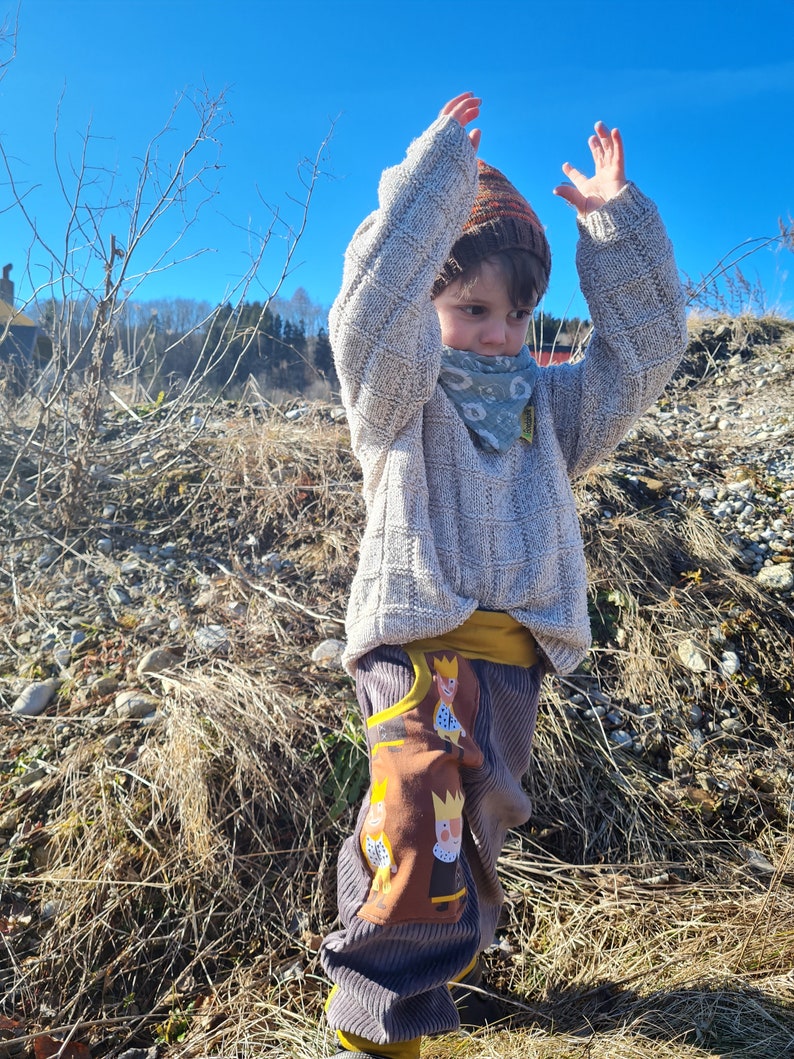 May include: A child wearing a light brown sweater with a white knit pattern, a brown and orange knit hat, and a green bandana with a white floral pattern. The child is wearing brown corduroy pants with a yellow waistband and a brown patch with a cartoon design of three people wearing crowns.