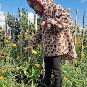 Pode incluir: Casaco com capuz com estampa de leopardo bege e preto com acabamento rosa. O casaco é usado com calças pretas e sapatos pretos. O fundo inclui uma cerca de madeira, flores amarelas e um céu azul.