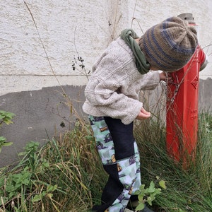 May include: A child wearing a beige sweater, a striped knit hat, and black pants with a green and white truck pattern. The child is standing next to a red fire hydrant.