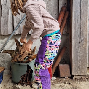Op de afbeelding: Een jong persoon draagt een lichtbruine trui en een paarse corduroy broek met een kleurrijke bloemenprint. De broek heeft een opgestikte zak op het rechterbeen. De persoon draagt witte sneakers met roze accenten.