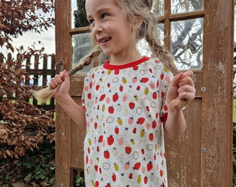 May include: A young girl wearing a white t-shirt with a red trim and a strawberry print. The shirt has two pockets on the sides. The girl has her hair in two braids and is smiling.