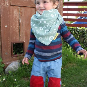 Puede incluir: Un ni&ntilde;o peque&ntilde;o con una camisa de manga larga a rayas azules y rojas con un pa&ntilde;uelo verde con estampados de animales blancos. El pa&ntilde;uelo tiene el texto "Boogie Bear" en la esquina inferior derecha. El ni&ntilde;o lleva pantalones vaqueros azules con rodilleras rojas y est&aacute; de pie sobre una superficie cubierta de hierba.