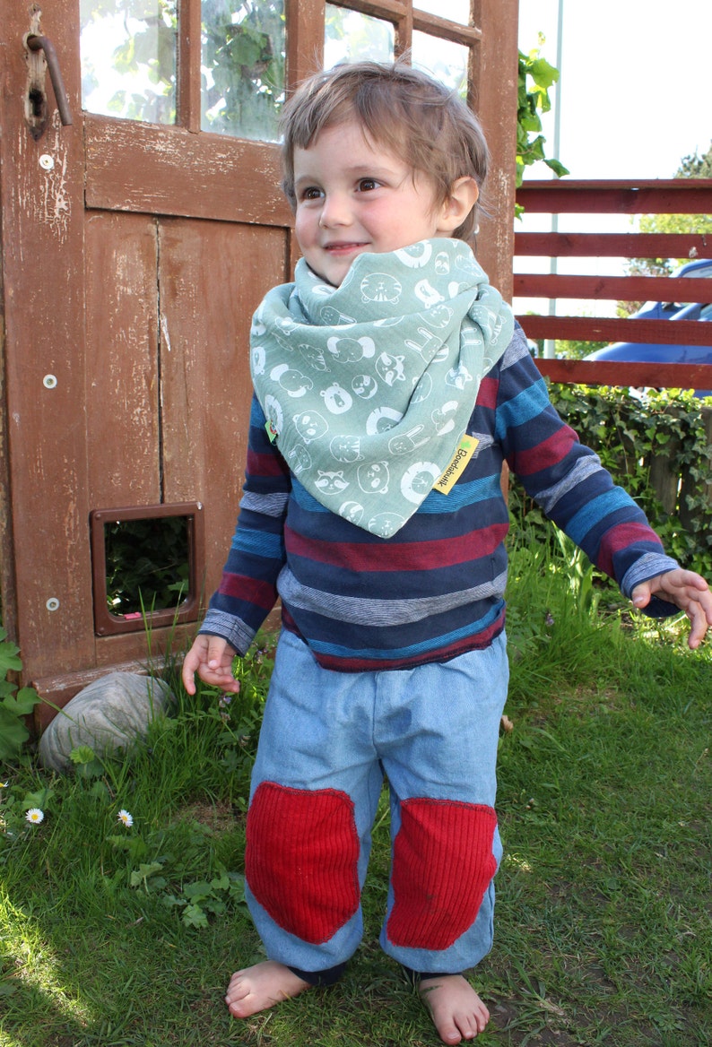 Puede incluir: Un ni&ntilde;o peque&ntilde;o con una camisa de manga larga a rayas azules y rojas con un pa&ntilde;uelo verde con estampados de animales blancos. El pa&ntilde;uelo tiene el texto "Boogie Bear" en la esquina inferior derecha. El ni&ntilde;o lleva pantalones vaqueros azules con rodilleras rojas y est&aacute; de pie sobre una superficie cubierta de hierba.