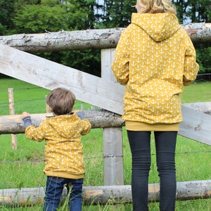 May include: A yellow hooded sweatshirt with a white anchor pattern. A child and an adult are wearing the sweatshirt. The adult is wearing black leggings and white sneakers. The child is wearing blue jeans and white sneakers. The two are standing in front of a wooden fence.
