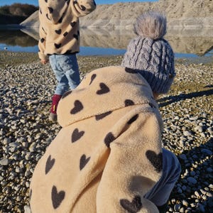 Puede incluir: Dos niños con chaquetas de forro polar de color crema con estampados de corazones negros. El niño en primer plano lleva un gorro de punto gris con un pompón esponjoso. Los niños están de pie en una playa de guijarros.