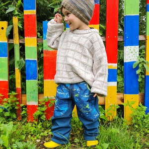 May include: A young child wearing a light brown knit sweater and blue jeans with a shark print stands in front of a colorful wooden fence. The child is wearing a striped knit hat and is smiling.