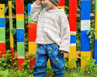 May include: A young child wearing a light brown knit sweater and blue jeans with a shark print stands in front of a colorful wooden fence. The child is wearing a striped knit hat and is smiling.