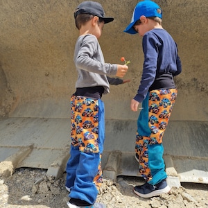 Op de afbeelding: Twee kinderen dragen blauwe en oranje broeken met een monstertruckprint en staan voor een groot stuk bouwmaterieel. Het kind links draagt een grijs shirt en een zwarte baseballpet. Het kind rechts draagt een blauw shirt en een blauwe baseballpet.