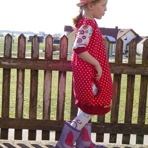 Puede incluir: Una ni&ntilde;a peque&ntilde;a con un vestido de lunares rojos y blancos con estampado floral en las mangas. Lleva un sombrero de paja con una cinta roja y botas de lluvia moradas. Est&aacute; de pie frente a una valla de madera.