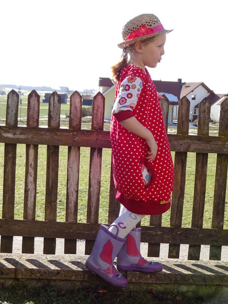 Puede incluir: Una ni&ntilde;a peque&ntilde;a con un vestido de lunares rojos y blancos con estampado floral en las mangas. Lleva un sombrero de paja con una cinta roja y botas de lluvia moradas. Est&aacute; de pie frente a una valla de madera.