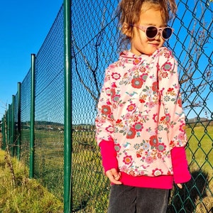 May include: A young person wearing a pink hoodie with a floral and bird print, black pants, and sunglasses. The person is standing in front of a green chain link fence.