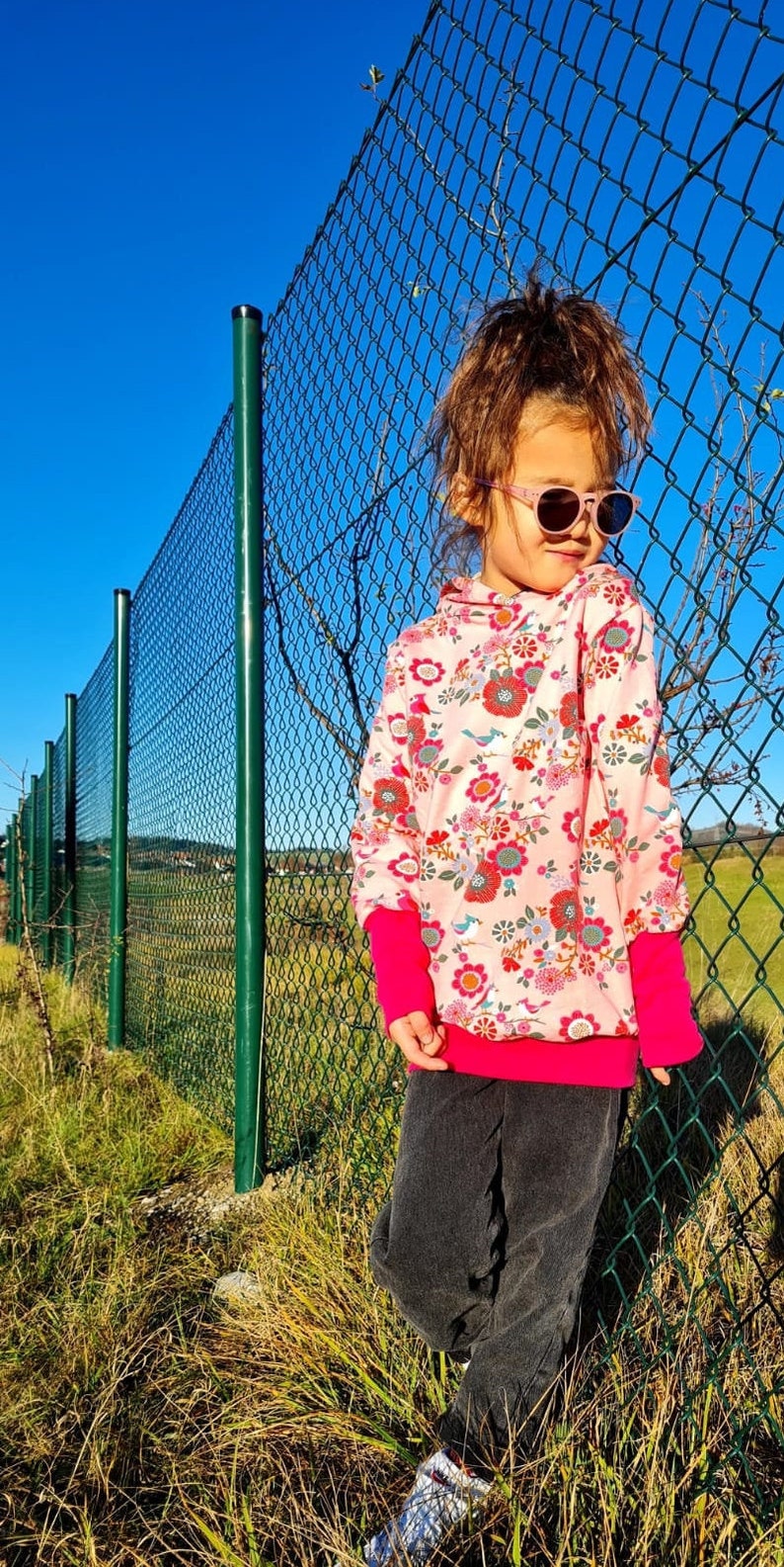 May include: A young person wearing a pink hoodie with a floral and bird print, black pants, and sunglasses. The person is standing in front of a green chain link fence.