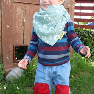 Puede incluir: Un ni&ntilde;o peque&ntilde;o con una camisa de manga larga a rayas azules y rojas con un pa&ntilde;uelo verde con estampado de animales blancos. El pa&ntilde;uelo tiene el texto "Goodthink". El ni&ntilde;o lleva pantalones vaqueros azules con rodilleras rojas y est&aacute; descalzo. El ni&ntilde;o est&aacute; de pie frente a una puerta de madera con una ventana.