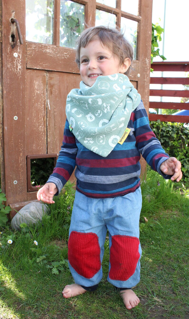 Puede incluir: Un ni&ntilde;o peque&ntilde;o con una camisa de manga larga a rayas azules y rojas con un pa&ntilde;uelo verde con estampado de animales blancos. El pa&ntilde;uelo tiene el texto "Goodthink". El ni&ntilde;o lleva pantalones vaqueros azules con rodilleras rojas y est&aacute; descalzo. El ni&ntilde;o est&aacute; de pie frente a una puerta de madera con una ventana.