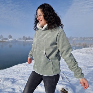 Könnte beinhalten: Eine Frau trägt einen hellgrünen Kapuzenpullover mit weißem Fleecefutter, schwarze Jeans und eine Brille. Sie steht auf einem schneebedeckten Feld mit einem Gewässer im Hintergrund.