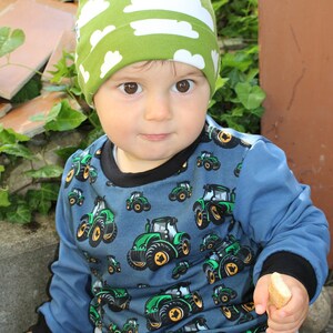 May include: A young child wearing a green and white hat with a cloud pattern and a blue long-sleeved shirt with a tractor pattern. The child is holding a piece of bread in their right hand.
