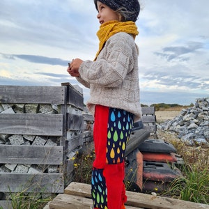 Op de afbeelding: Een jong kind draagt een rode broek met een regenboog- en regendruppelpatroon. De broek heeft een zwart-witte streep aan de zijkant. Het kind draagt ook een beige trui en een gele sjaal.