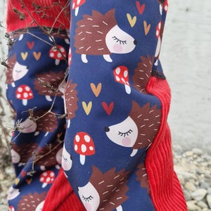 Op de afbeelding: Rode corduroy broek met blauwe stofpanelen met een herhalend patroon van bruine egels met gesloten ogen en rode en witte paddenstoelen. De stofpanelen hebben een wit hartpatroon met gele en oranje accenten.