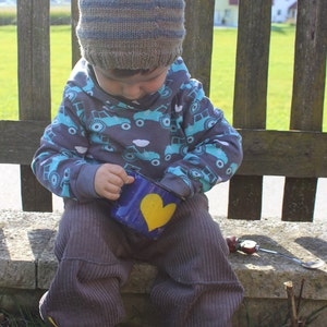 May include: A young child wearing a brown and blue striped knitted hat, a grey sweater with a blue and white truck pattern, and brown corduroy trousers. The child is holding a blue cup with a yellow heart on it.