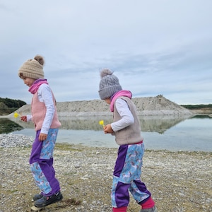 Op de afbeelding: Twee kinderen dragen bijpassende paarse en blauwe broeken met een eenhoornprint, roze en grijze vesten en mutsen en lopen op een rotsachtig pad in de buurt van een waterpartij.
