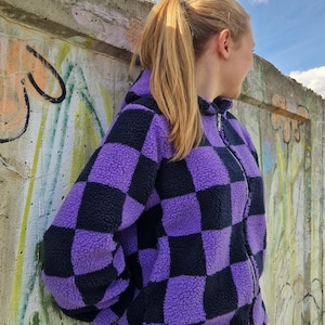 Könnte beinhalten: Eine langärmelige Reißverschlussjacke aus flauschigem, strukturiertem Fleece-Material mit einem auffälligen, sich wiederholenden Schachbrettmuster aus abwechselnden Blöcken in tiefem Schwarz und leuchtendem Violett. Die Jacke hat einen Stehkragen.