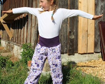 May include: A young person wearing a white long-sleeved shirt and purple patterned pants. The pants have a wide purple waistband and a light purple background with peacock and paisley designs. The person is standing on green grass.