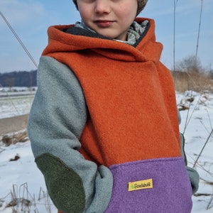 May include: A child wearing a multi-coloured hooded sweatshirt with a green, blue, orange and purple colour scheme. The sweatshirt has a small yellow label with the text "Boedabuitik" on the front. The child is wearing a brown and blue knitted hat.