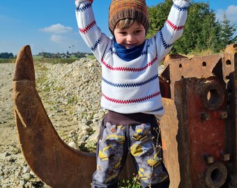 May include: A young child wearing a white and red striped sweater, a brown and orange knit hat, and blue pants with a construction vehicle pattern. The child is standing in a gravel pit with a large rusty metal object behind them.