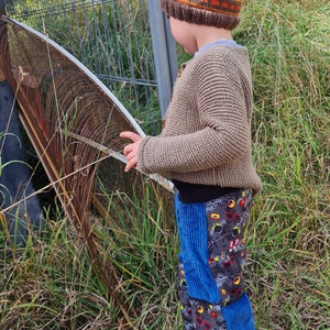 Op de afbeelding: Een kind draagt ​​een bruine en oranje gebreide muts met een geometrisch patroon, een bruine gebreide trui en een blauwe corduroy broek met een kleurrijke grafische print. Het kind staat in een grasachtig gebied in de buurt van een metalen hek.