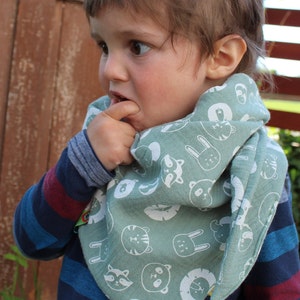 Puede incluir: Un ni&ntilde;o peque&ntilde;o con una camisa de manga larga a rayas azules y rojas con un pa&ntilde;uelo verde con ilustraciones de animales blancos. El pa&ntilde;uelo tiene una variedad de caras de animales, incluyendo un mapache, un oso, un tigre, un conejo y un le&oacute;n.