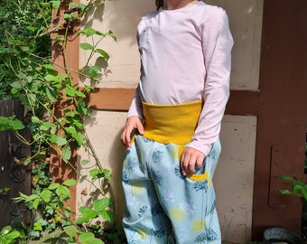 May include: A young child wearing blue pants with a white and yellow floral pattern, a yellow waistband, and a pink long-sleeved shirt. The child is standing on a rock in a garden setting.