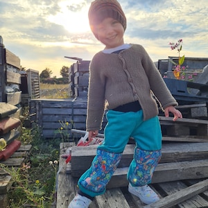 Op de afbeelding: Een jong kind draagt ​​een bruin gebreid vest, een bruin en oranje gebreide muts en een blauwe corduroy broek met een kleurrijk printje. Het kind staat op een houten pallet buiten.
