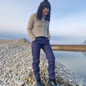 Op de afbeelding: Een persoon draagt een lichtbruine trui en een donkerblauwe broek en staat op een bed van stenen aan de rand van een waterlichaam.