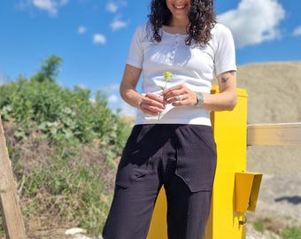 Puede incluir: Una persona lleva una camiseta blanca de manga corta y pantalones negros. La persona sostiene una flor amarilla. El fondo incluye un cielo azul brillante con nubes y una estructura amarilla.