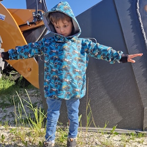 May include: A young person wearing a blue hooded sweatshirt with a monster truck print. The sweatshirt has a black hood and black cuffs. The person is wearing blue jeans and black shoes.