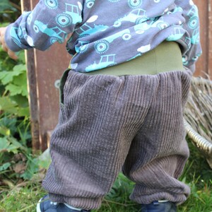 May include: A child wearing brown corduroy trousers with a green waistband and a blue and turquoise patterned long-sleeved shirt with a hood.