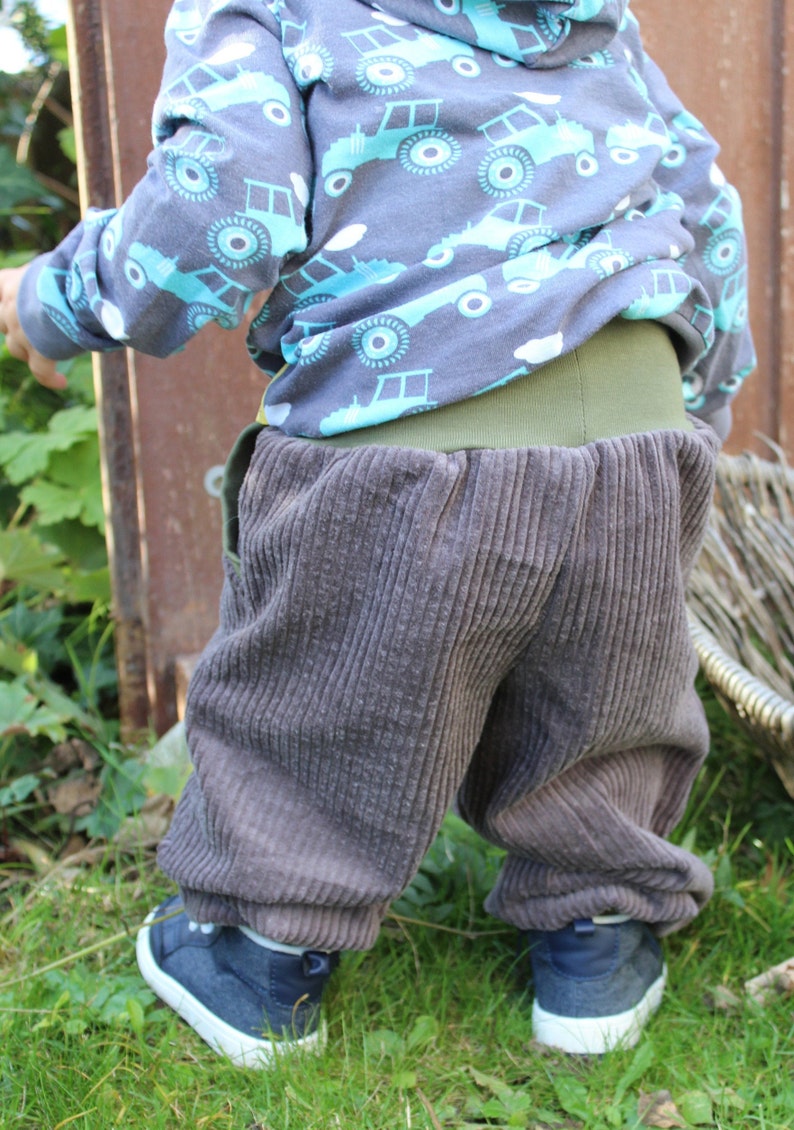 May include: A child wearing brown corduroy trousers with a green waistband and a blue and turquoise patterned long-sleeved shirt with a hood.