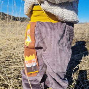 May include: Gray corduroy pants with a yellow waistband and two embroidered patches of cartoon kings wearing crowns. The patches are on the front of the pants, one on each leg.