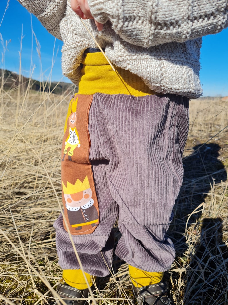 May include: Gray corduroy pants with a yellow waistband and two embroidered patches of cartoon kings wearing crowns. The patches are on the front of the pants, one on each leg.