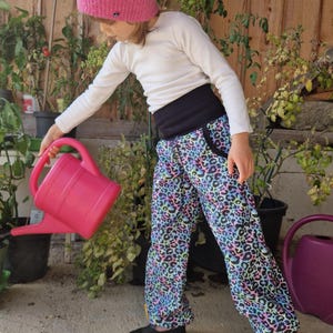 Puede incluir: Un niño con un gorro de punto rosa, una camiseta blanca de manga larga y pantalones estampados con zapatos negros. Los pantalones tienen una cintura negra y un estampado de leopardo colorido. El niño sostiene una regadera rosa.