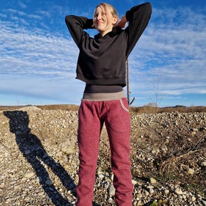 Op de afbeelding: Een jong persoon draagt een zwarte hoodie en een bordeauxrode fleecebroek met een grijze tailleband. Ze staan op een rotsachtig oppervlak met een blauwe lucht op de achtergrond.