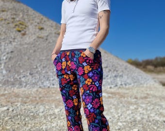 May include: A person wearing a white short-sleeved shirt and floral print pants. The pants feature a dark background with colorful flowers in shades of red, orange, purple, and blue. The person is standing outdoors on a rocky surface.
