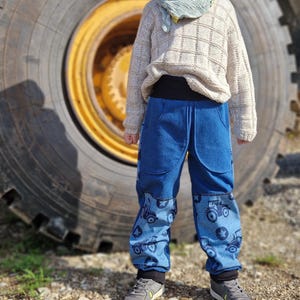 Op de afbeelding: Een jong persoon draagt een beige trui, een groene beanie en een blauwe sjaal. Ze dragen een blauwe jeans met een tractorprint en grijze sneakers. De persoon staat voor een grote band.