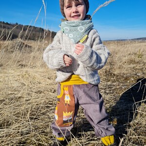 May include: A young child wearing a brown knitted hat, a light blue bandana, a white sweater, and grey corduroy pants with a brown pocket featuring a cartoon design. The child is holding a tall blade of grass in a field.