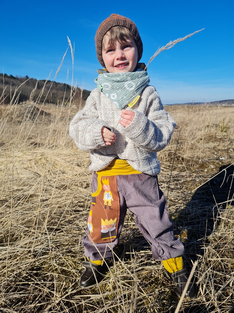 May include: A young child wearing a brown knitted hat, a light blue bandana, a white sweater, and grey corduroy pants with a brown pocket featuring a cartoon design. The child is holding a tall blade of grass in a field.